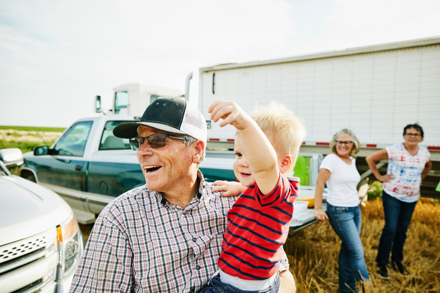 Medium shot of laughing farmer holding grandson during family meal in wheat field during harvest