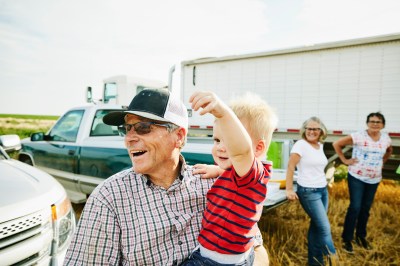 Medium shot of laughing farmer holding grandson during family meal in wheat field during harvest