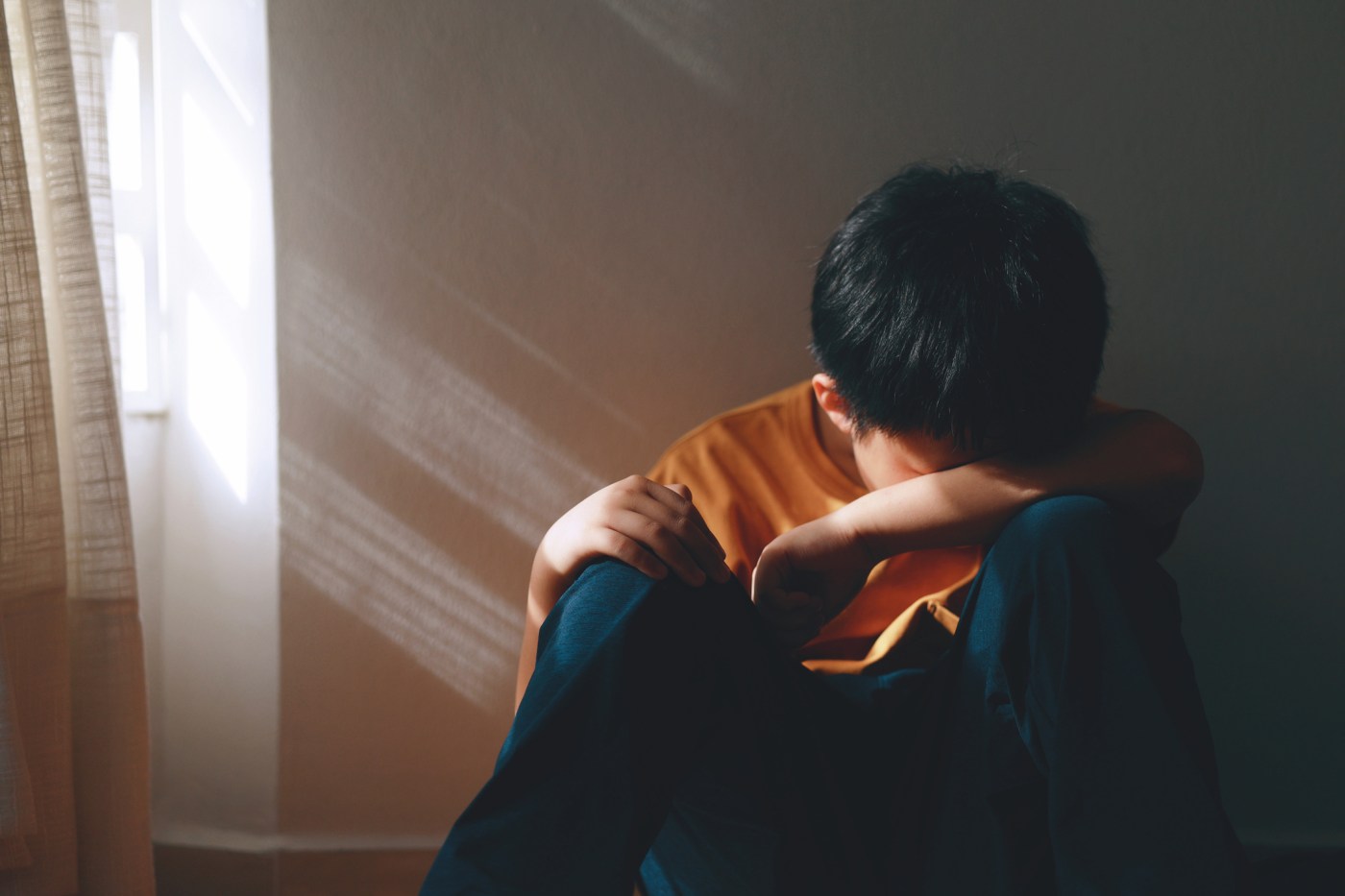Boy crying in a corner, his face is illuminated by the shadowy light coming through a window.