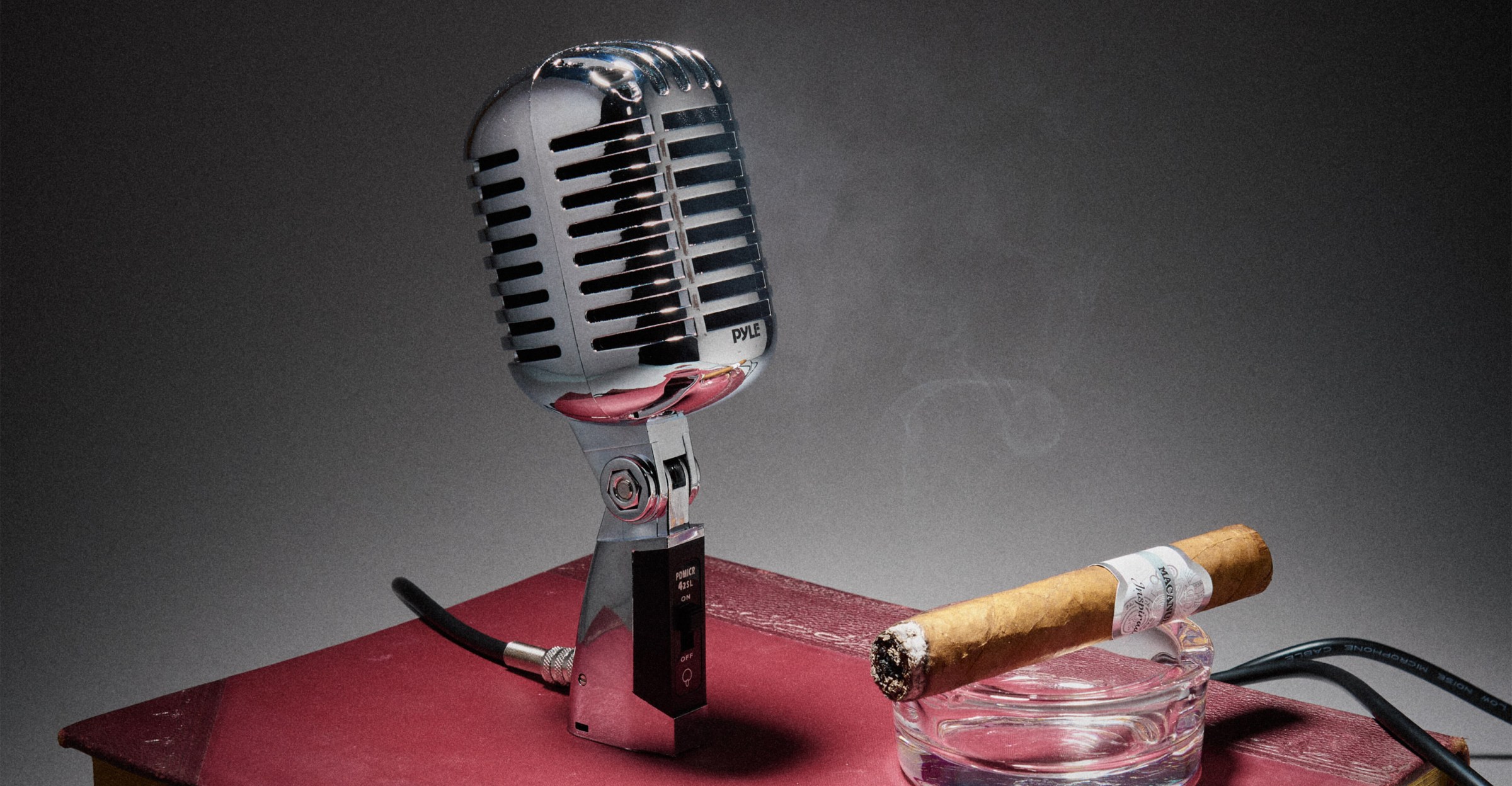 A vintage silver microphone stands on a red book beside a lit cigar in a glass ashtray emitting smoke, set against a moody, dimly lit background.